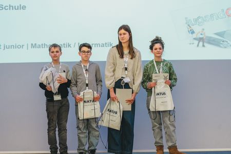 Vier Kinder und Jugendliche stehen nebeneinander auf einer Bühne und halten Urkunden sowie Stofftaschen bei der Preisverleihung von ‚Jugend forscht‘.