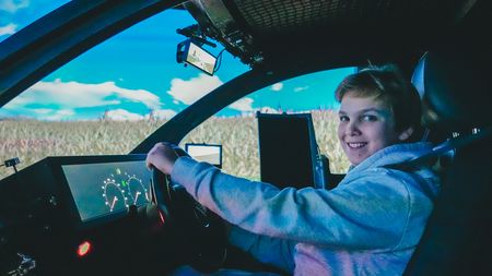 Kind sitzt am Steuer eines Fahrzeugsimulators und lächelt in die Kamera; vor ihm sind digitale Anzeigen und Bildschirme zu sehen, im Hintergrund eine projizierte Landschaft.
