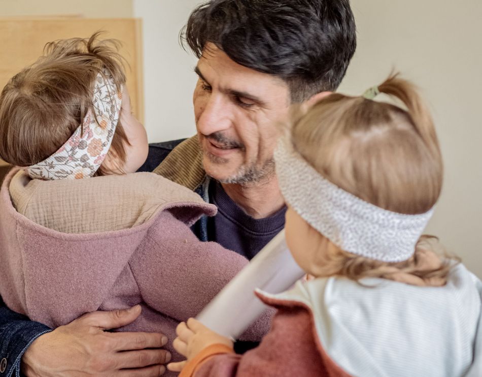 Ein Vater hält seine zwei kleinen Kinder auf dem Arm und lächelt ihnen zu. Die Kinder tragen Stirnbänder und sind ihm zugewandt.
